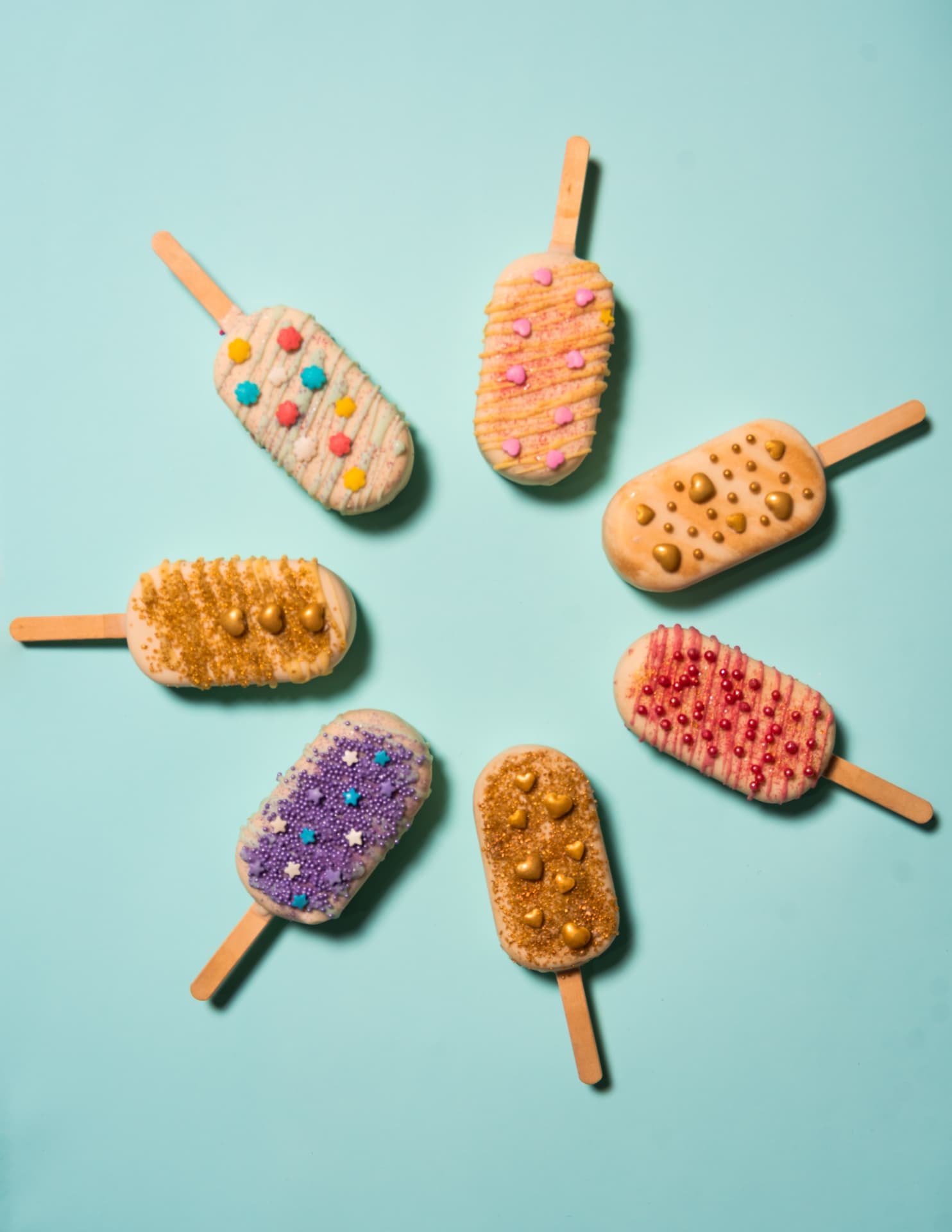 Vista cenital de siete helados palito decorados con confites, dispuestos en círculo sobre fondo turquesa