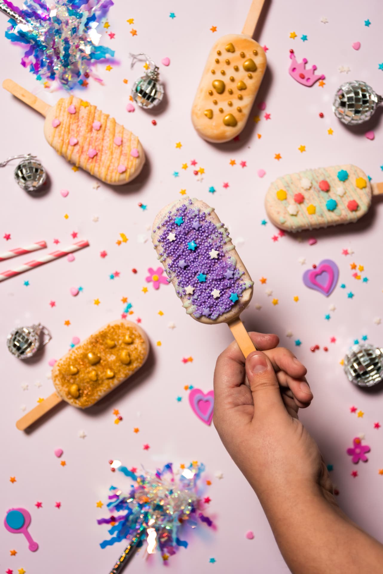 Vista cenital de helados palito decorados con confites, bolas de espejos y corazones sobre fondo rosa pálido