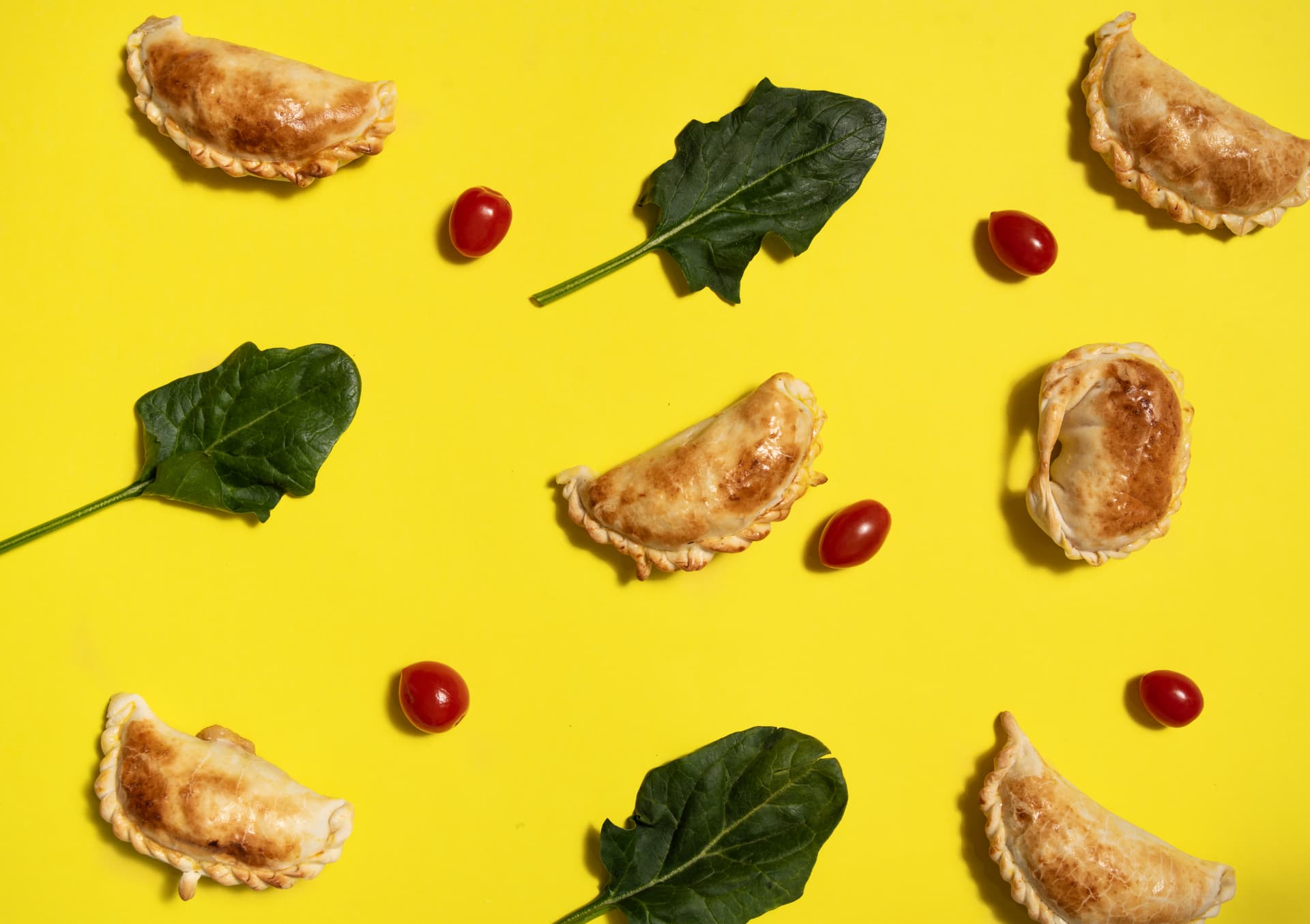 Composición cenital con empanadas, hojas verdes y tomates sobre fondo amarillo