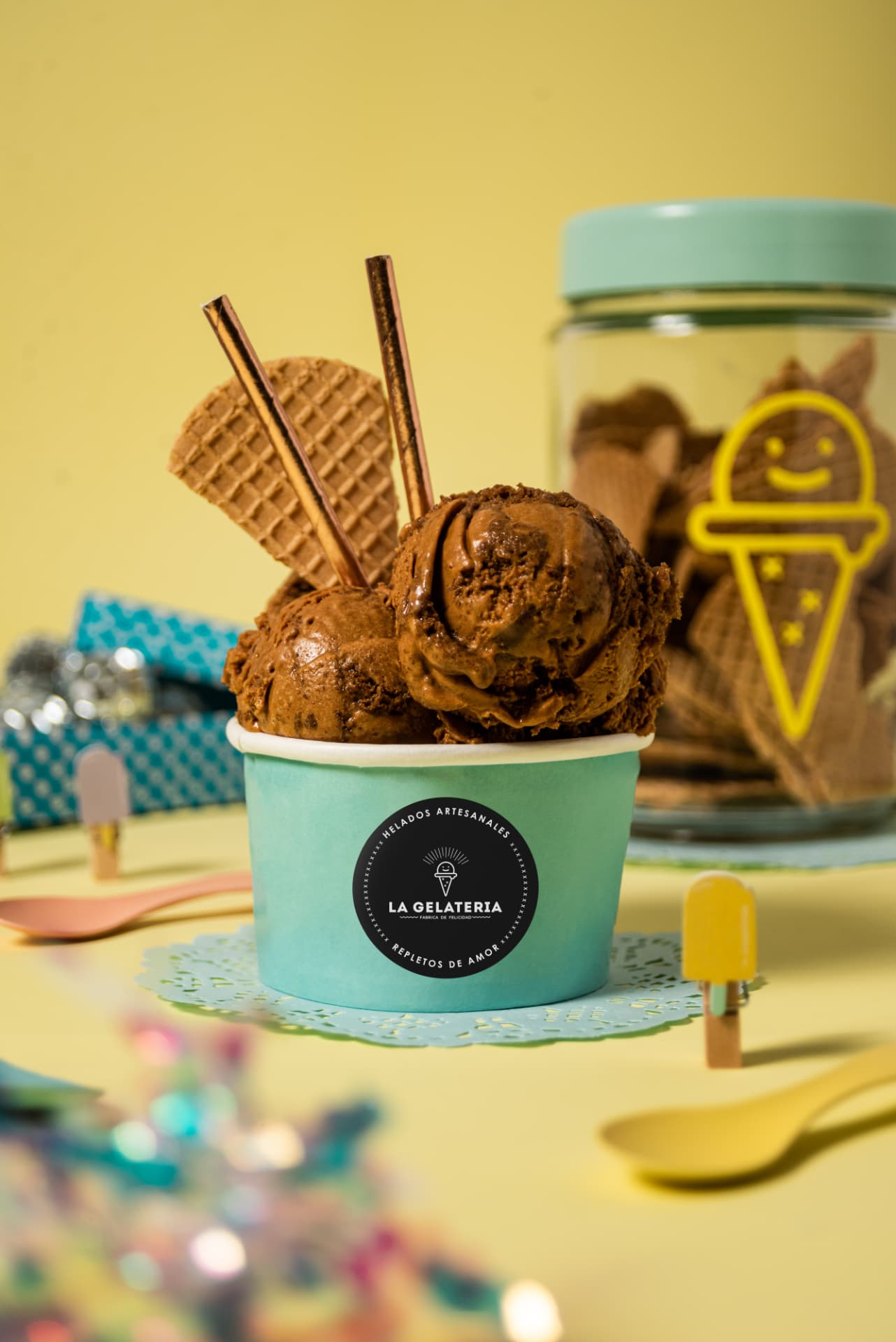 Pote de helado de chocolate con barquillo y sorbetes cobre sobre fondo amarillo, frasco de barquillos detrás