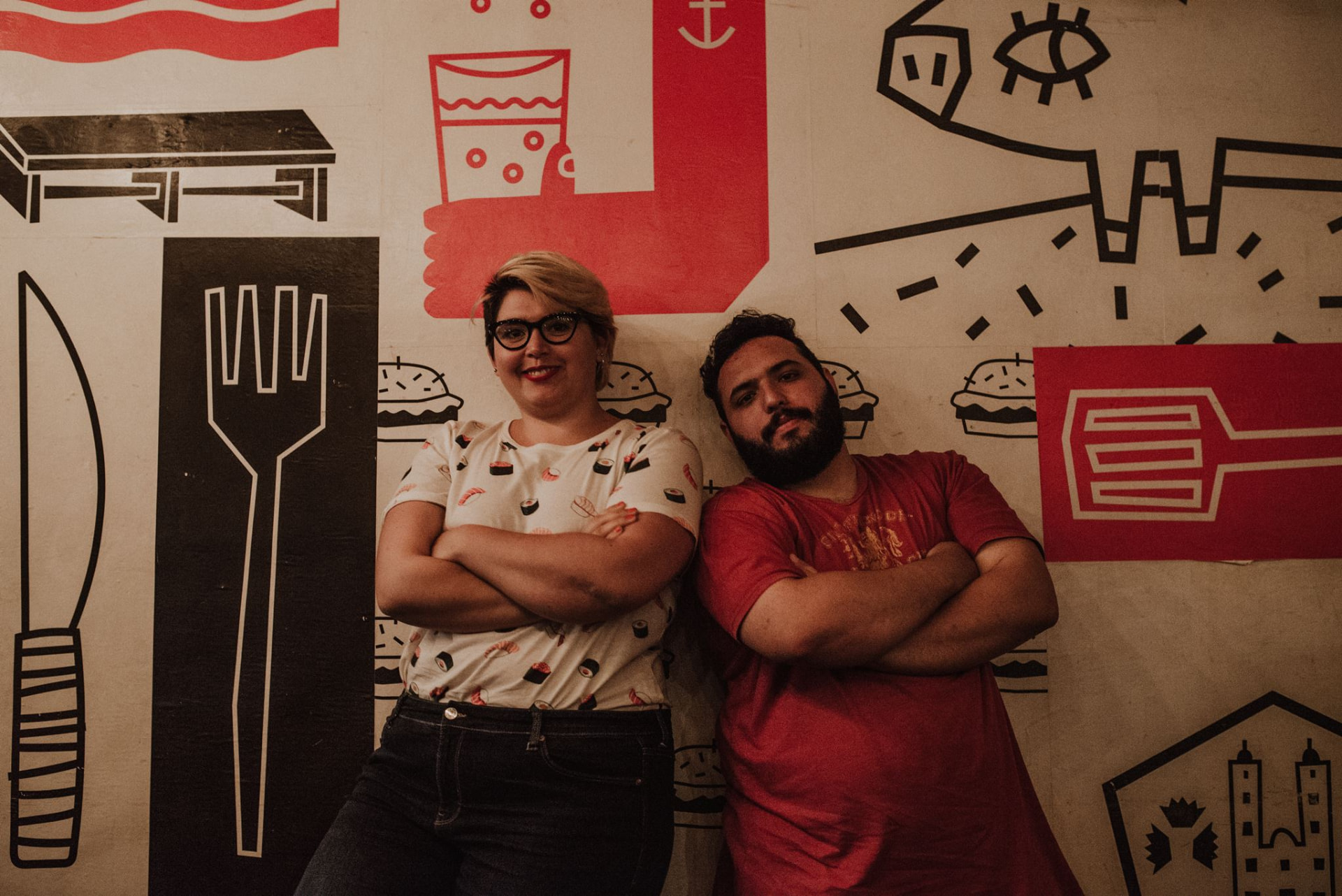 Gabriela Gaona y Carma Pelliccia de pie, con los brazos cruzados, frente a una pared ilustrada con piezas gráficas gastronómicas