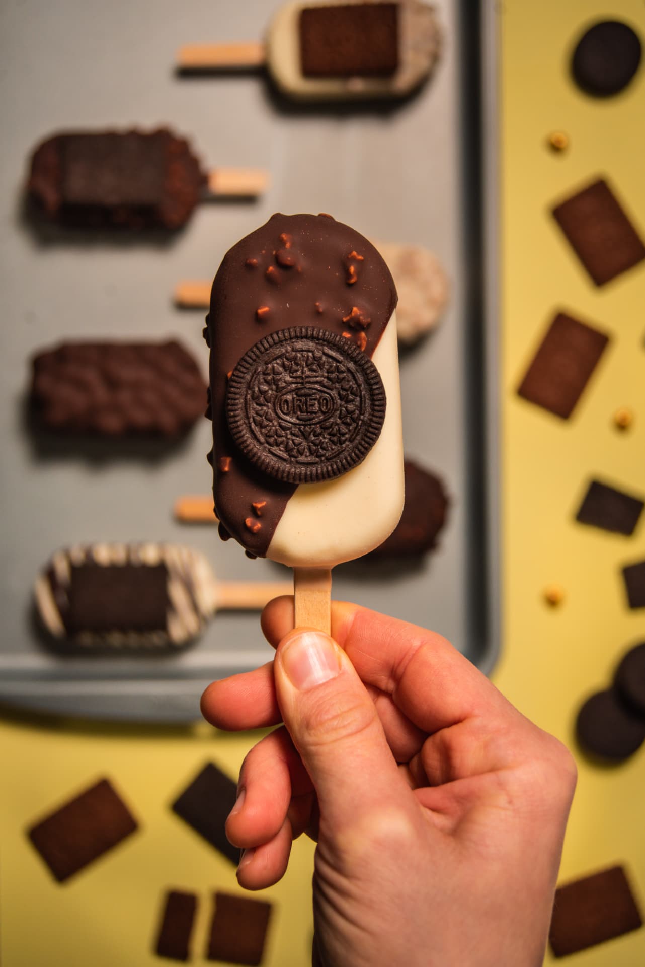 Mano sosteniendo un helado palito bañado en chocolate con galletita, bandeja de palitos y galletitas sobre fondo amarillo